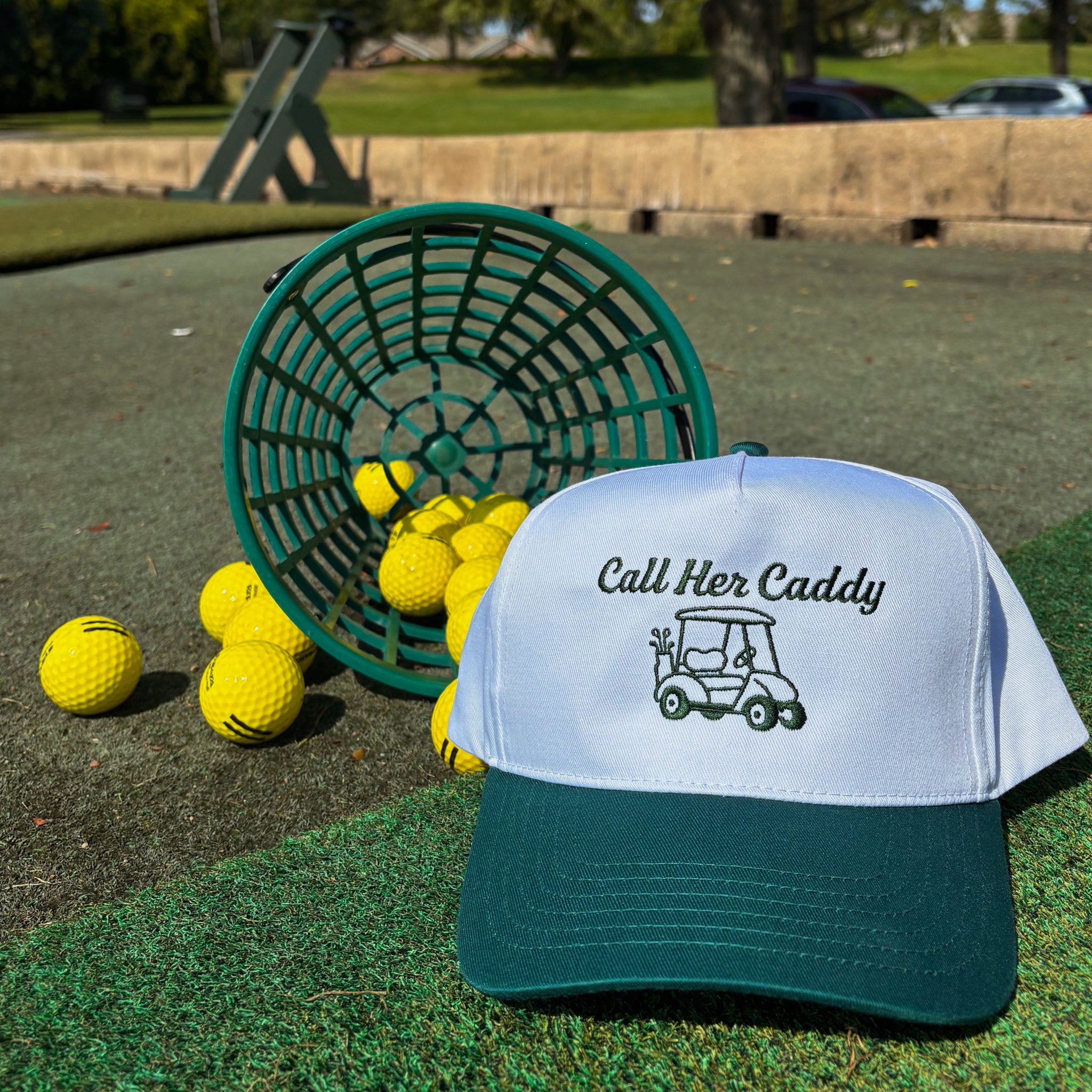 Golf hat with 'Call Her Caddy' text and design on a grassy area with golf balls and a basket.
