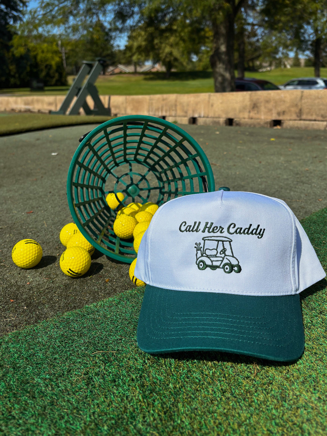 Golf hat with 'Call Her Caddy' text and design on a grassy area with golf balls and a basket.