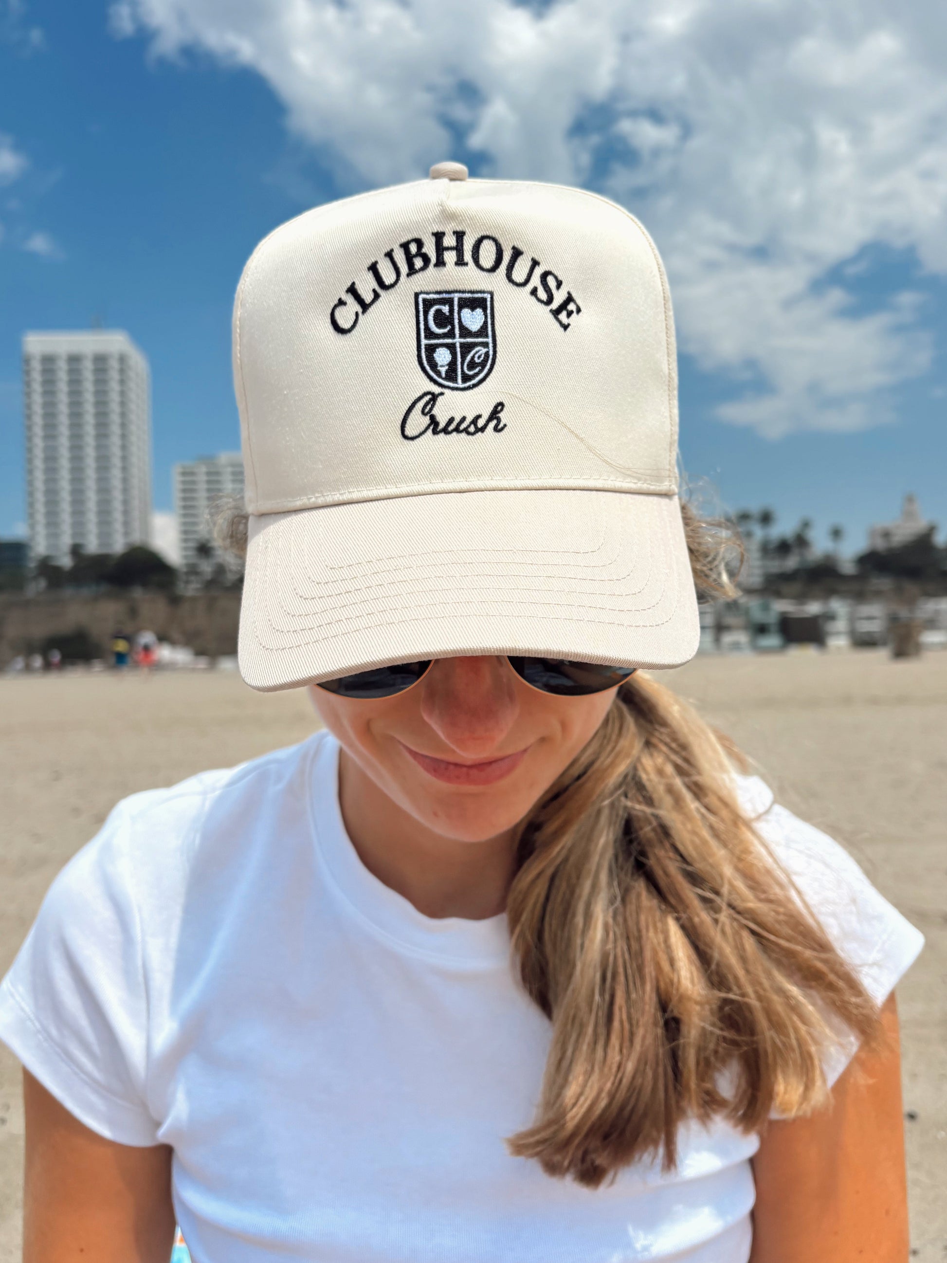 Person wearing a beige cap with 'Clubhouse Crush' text on a beach